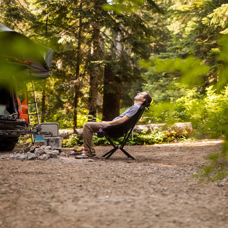 Person relaxing in a CLIQ EpiQ Lounge Chair at a campsite with trees and camping gear in the background