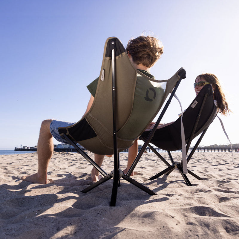 Two people sitting on CLIQ MystiQ portable beach chairs on a sandy beach with a clear sky.