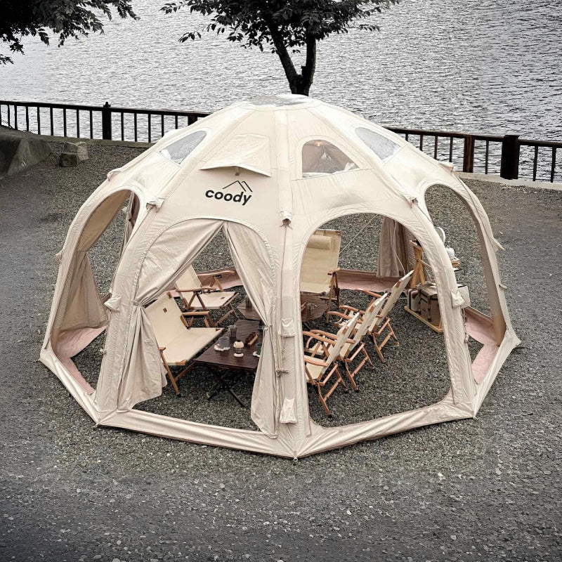 Coody Aurora Dome inflatable tent in beige with furniture inside, set up near a body of water.