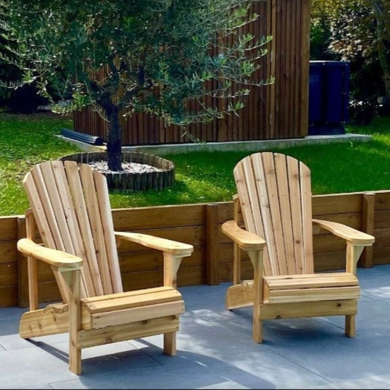 Two wooden Adirondack chairs by The Best Adirondack Chair Company on a patio with a garden and tree in the background.