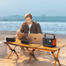 Man using a laptop on a beach with a Jackery portable power station powering devices.