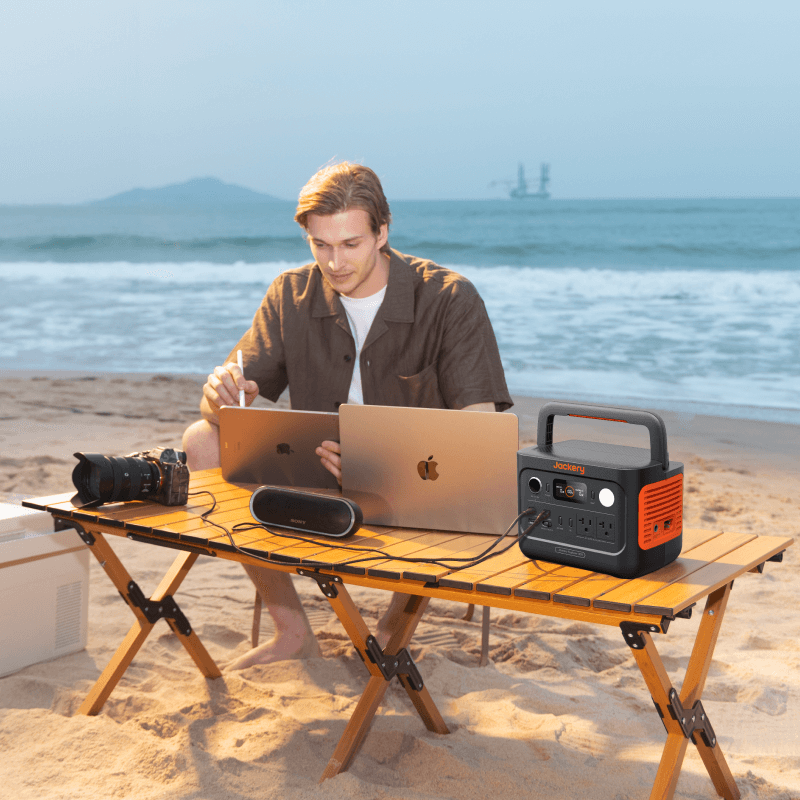 Man using a laptop on a beach with a Jackery portable power station powering devices.